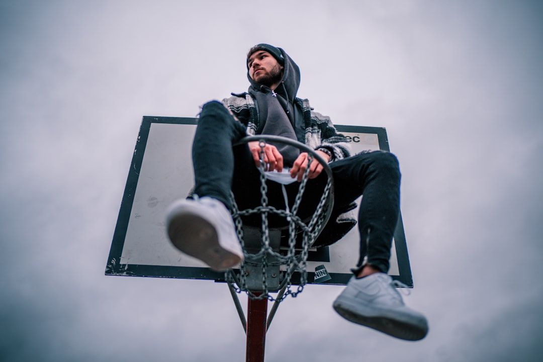 Eine Person mit Kapuzenjacke und Beanie sitzt auf einem Basketballkorb und hält den Ring mit beiden Händen fest. Der Himmel über Aachen ist bedeckt.