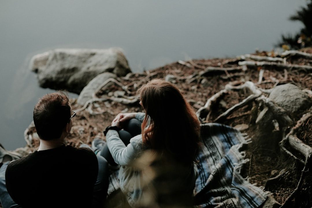 Zwei Menschen sitzen auf einer Decke neben einem ruhigen Gewässer, umgeben von Felsen und Baumwurzeln, mit dem Rücken zur Kamera. Dies erinnert an eine ruhige Szene am Stadtrand von Aachen.