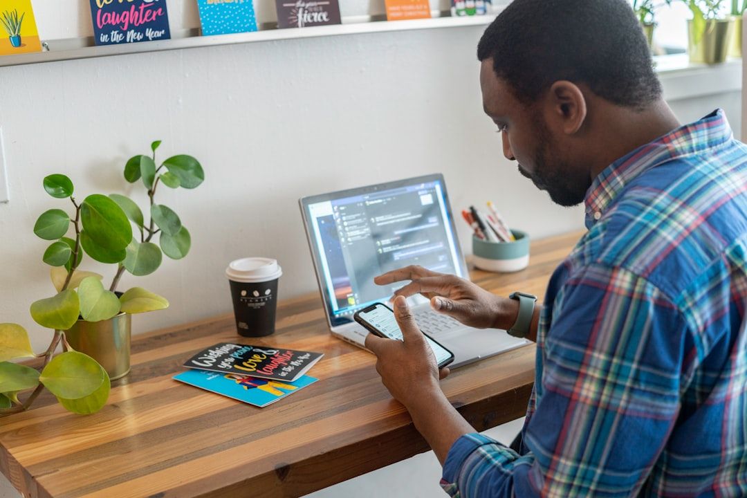 Ein Mann sitzt an einem Holzschreibtisch in Aachen und arbeitet an Laptop und Telefon. Auf dem Schreibtisch stehen außerdem eine Pflanze, eine Kaffeetasse und ein Buch mit dem Titel „Strategien“.