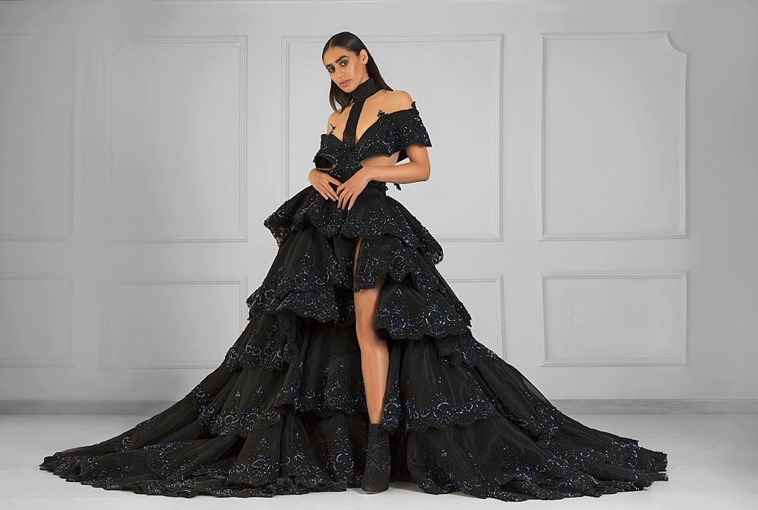 Eine Frau in einem bodenlangen, schwarzen Lagenkleid mit schulterfreien Ärmeln und aufwendigen Details steht mit einer Eleganz, die an Aachens historischen Charme erinnert, vor einer schlichten, grauen Wand.