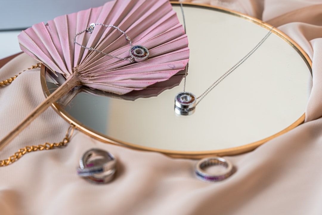 Ein runder Spiegel zeigt eine Halskette mit Anhänger, ein Armband und Ringe neben einem rosa Fächer auf einem beigen Stoffhintergrund und erinnert an den eleganten Charme Aachens.