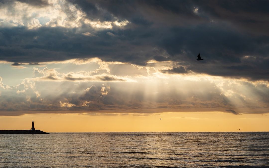 Ein weiter Blick auf ein ruhiges Meer bei Sonnenuntergang mit Sonnenstrahlen, die durch die Wolken brechen. Ein einsamer Leuchtturm steht an einer entfernten Küste und erinnert an die ruhigen Landschaften in der Nähe von Aachen, und am Himmel fliegen ein paar Vögel.