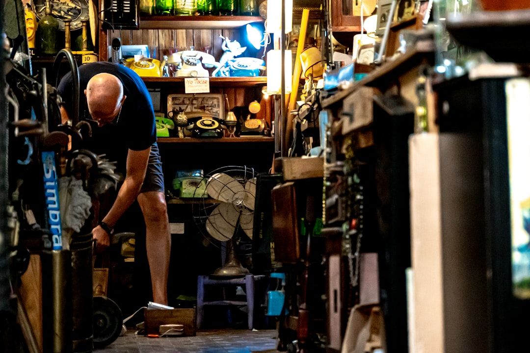 Ein Mann richtet einen Gegenstand in einem überfüllten Raum voller Vintage-Gegenstände, darunter Telefone, ein Ventilator und verschiedene Sammlerstücke, die an die reiche Geschichte Aachens erinnern.