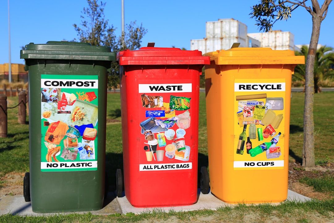 Drei farblich markierte Behälter mit den Aufschriften „Kompost – kein Plastik“, „Abfall – nur Plastiktüten“ und „Recycling – keine Becher“ stehen in einem Park in Aachen. Im Hintergrund sind Bäume und Industriegebäude zu sehen.