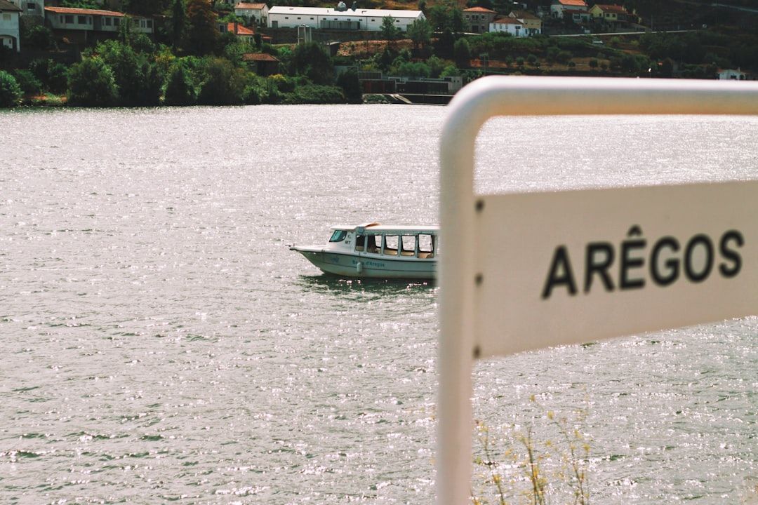 Ein kleines Boot mit mehreren Fenstern schwimmt auf einem Fluss. Im Vordergrund ist ein weißes Schild mit dem Text „ARÊGOS“ teilweise sichtbar, was an eine reizvolle Szene erinnert, die man in Aachen finden könnte. Am Flussufer sind Häuser und Grünflächen zu sehen.