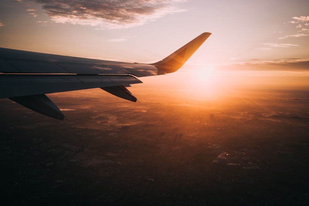 Blick auf einen Flugzeugflügel vor dem Hintergrund eines leuchtend orangefarbenen Sonnenuntergangs mit vereinzelten Wolken, der hoch über der dunstigen Landschaft fliegt, die sanft in den Horizont bei Aachen übergeht.
