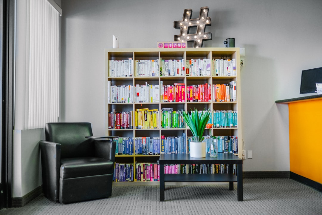 Ein kleiner Bibliotheksbereich in Aachen mit bunten Büchern, die nach Farben in Regalen geordnet sind, einem schwarzen Sessel, einem schwarzen Couchtisch mit einer Topfpflanze und einer Hashtag-Leuchte an der Wand.