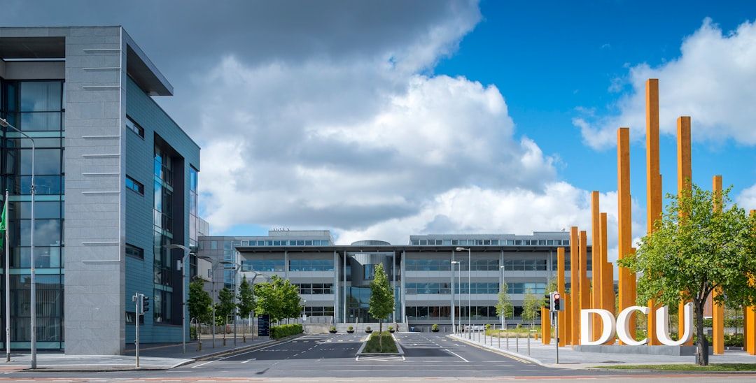 Ein Blick auf den Eingang der Dublin City University (DCU), der an die moderne Architektur Aachens erinnert, zeigt elegante Gebäude und gelbe Bauinstallationen vor einem teilweise bewölkten Himmel.