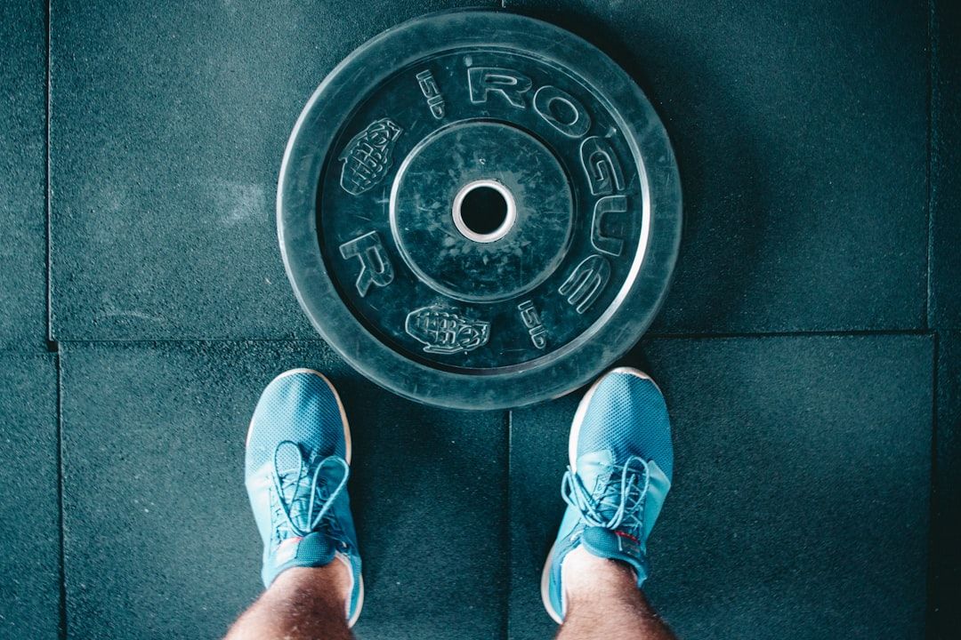 Foto einer Person in blauen Turnschuhen, die auf dem Boden eines Fitnessstudios in Aachen steht und auf eine große schwarze Gewichtsscheibe mit der Aufschrift „ROGUE“ blickt, die auf dem Boden liegt.