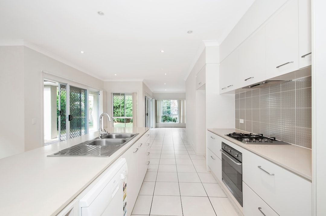 Moderne Küche mit weißen Schränken, Geräten aus Edelstahl und einer Doppelspüle. Der Raum verfügt über große Fenster und Glasschiebetüren, die zu einem Außenbereich mit viel Grün führen, der an die malerische Landschaft Aachens erinnert.