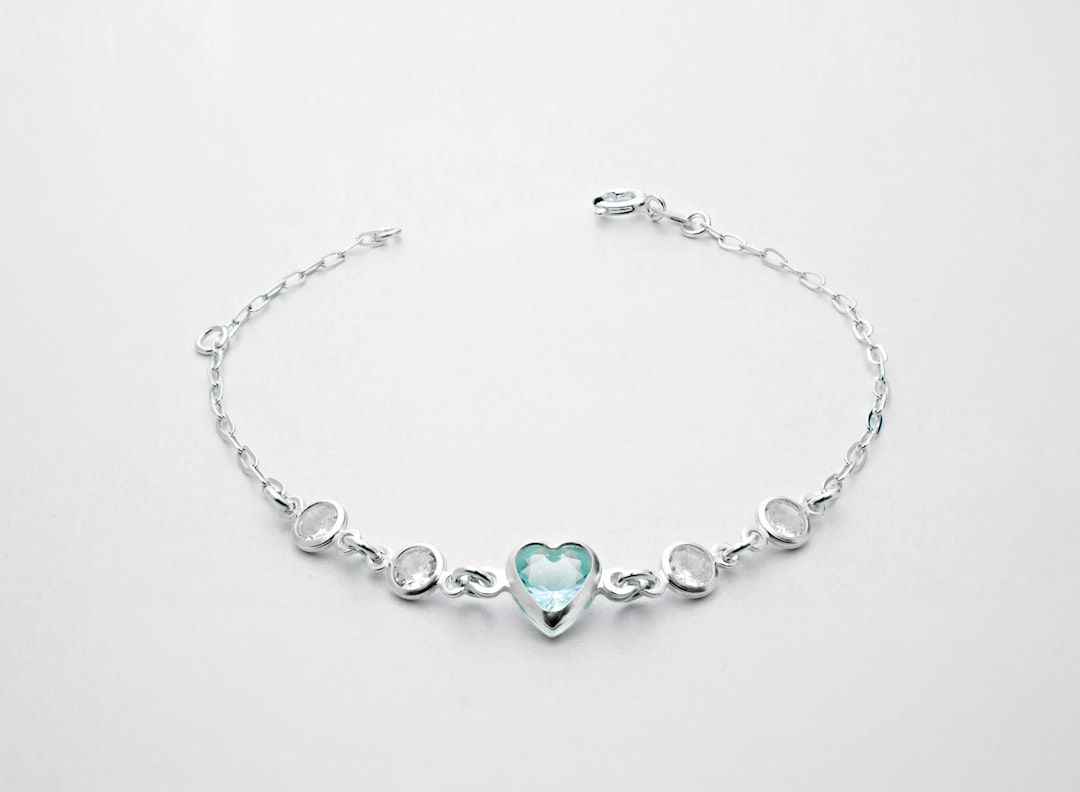 Ein silbernes Armband mit Kettengliedern, inspiriert vom historischen Charme Aachens. Es verfügt über ein herzförmiges Mittelstück aus blauen Edelsteinen und vier runde weiße Edelsteine.