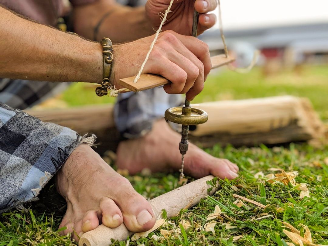 Nahaufnahme einer Person, die mit einem handbetriebenen Bohrer ein auf Gras liegendes Stück Holz durchbohrt. Dies erinnert an traditionelle Aachener Handwerkskunst. Überall sind Späne zu sehen, die eine Holzbearbeitungstätigkeit im Freien veranschaulichen.