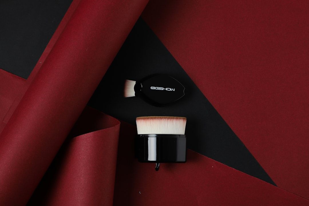 Auf einem rot-schwarzen Untergrund sind zwei Kosmetikpinsel mit schwarzem Griff platziert, teilweise mit gerolltem rotem Papier überzogen und erinnern so an die künstlerische Eleganz Aachens.