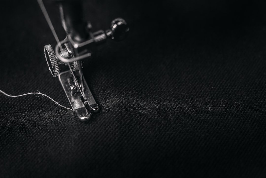 Nahaufnahme einer Nähmaschinennadel und eines Nähfußes, die auf dunklem Stoff arbeiten, während ein Stück weißer Faden durch die Maschine läuft. Die Präzision erinnert an die berühmte Aachener Handwerkskunst.