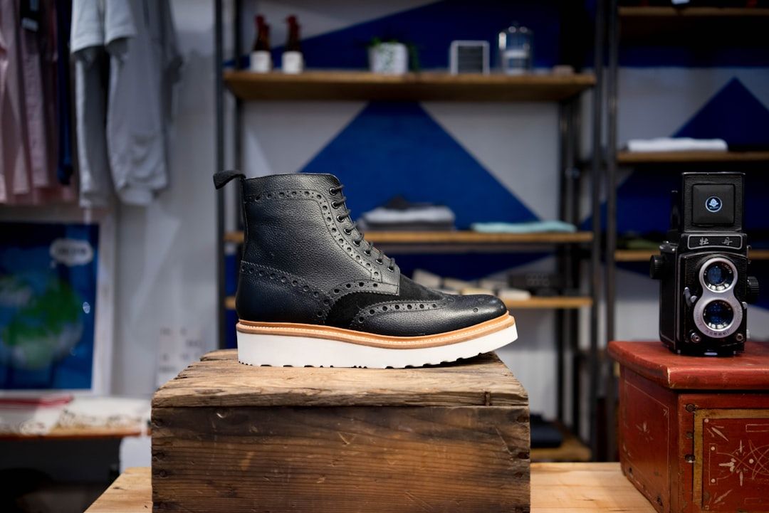 Ein schwarzer Leder-Brogue-Stiefel mit weißer Sohle steht auf einer rustikalen Holzkiste und erinnert an den klassischen Aachener Charme. Im Hintergrund sind Regale mit verschiedenen Artikeln zu sehen.