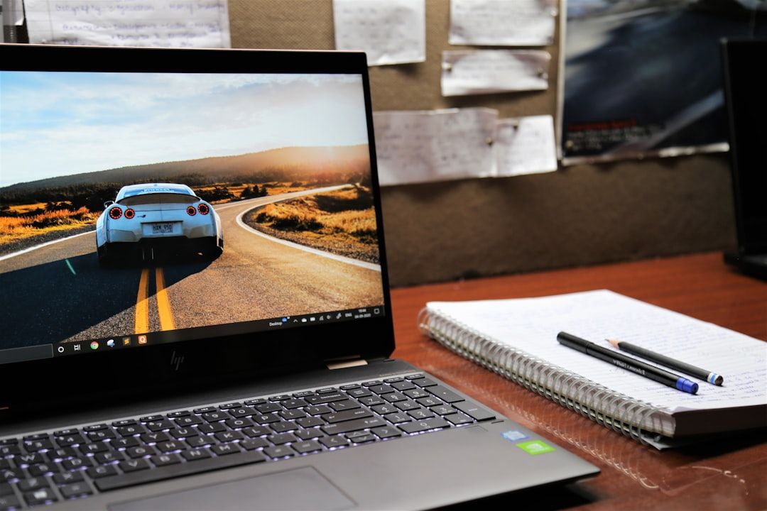 Auf einem Schreibtisch stehen ein Laptop mit einer Autotapete und ein Notizbuch mit Stiften, im Hintergrund sind Zettel mit Pinnwänden und Bilder aus Aachen an der Wand angeheftet.