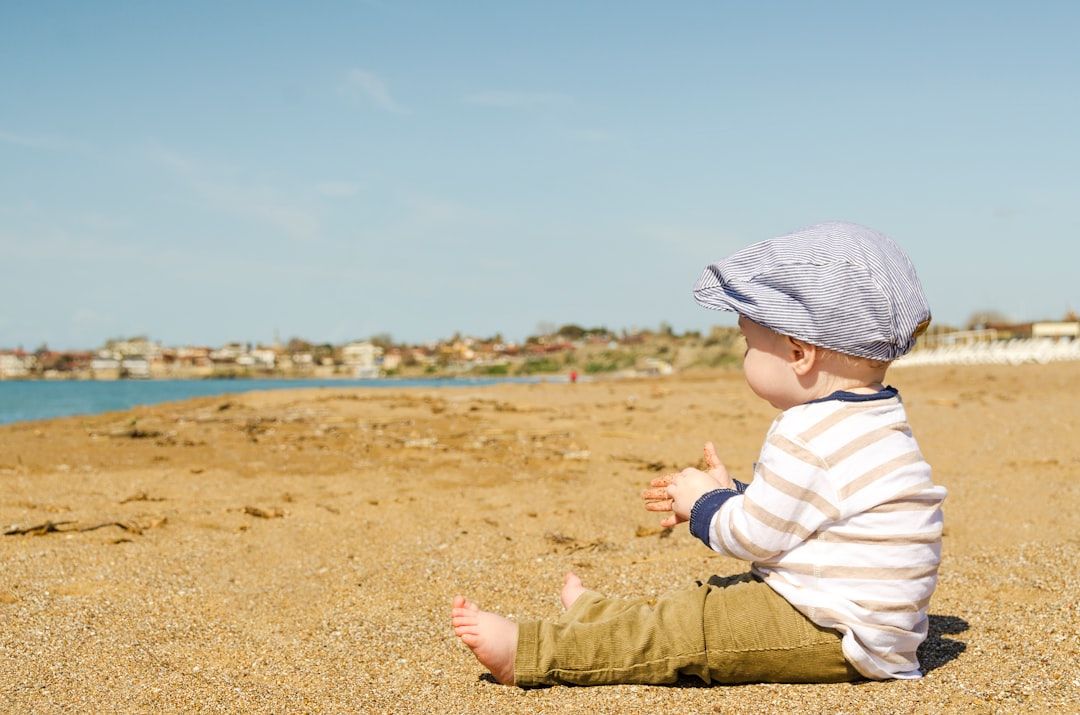 Ein Baby sitzt in einem gestreiften Hemd, Khakihosen und einer Mütze an einem Sandstrand und blickt auf das Wasser und die in der Ferne gelegenen Gebäude, die an die in Aachen erinnern.