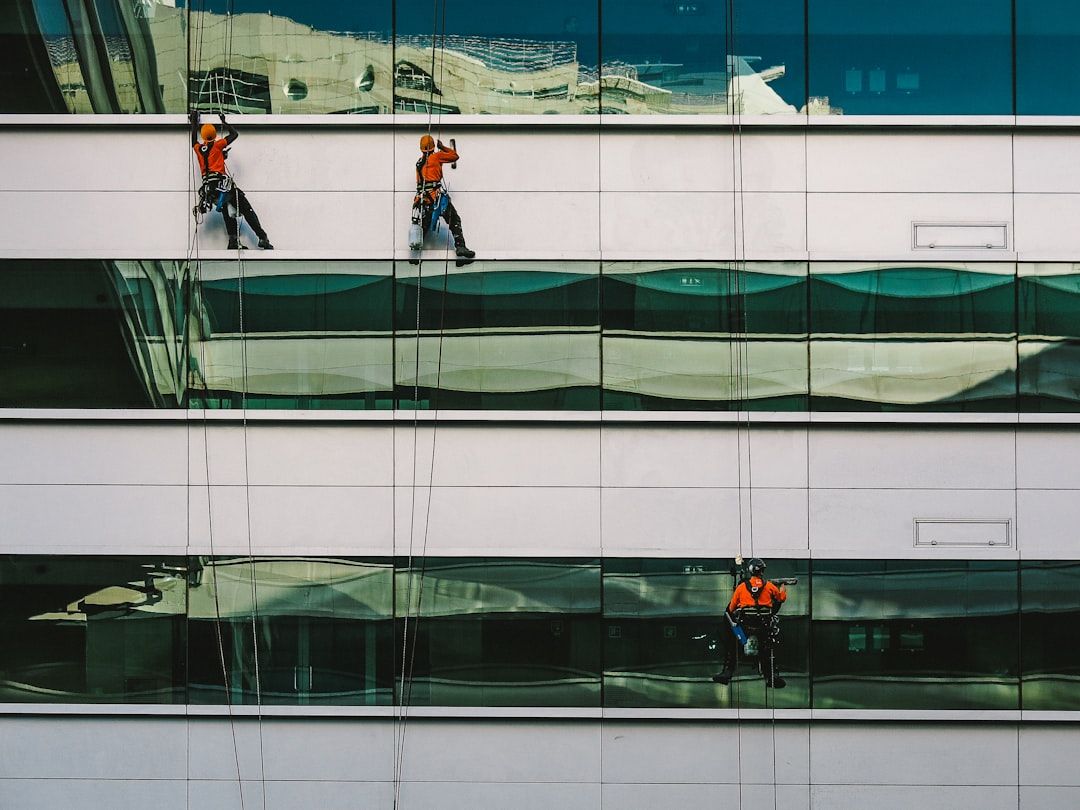 Drei Arbeiter in orangefarbenen Sicherheitswesten hängen an Seilen und putzen sorgfältig die Fenster eines Hochhauses in Aachen.