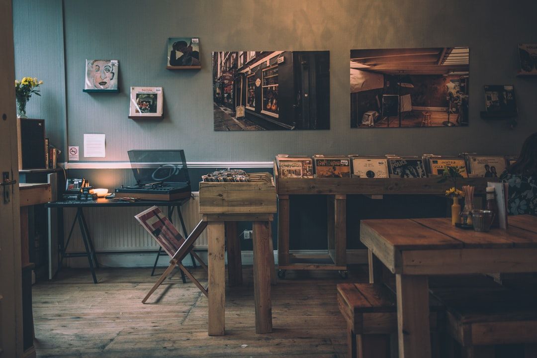 Ein gemütlicher Raum mit Holztischen und Regalen, auf denen Schallplatten und gerahmte Kunst ausgestellt sind. Auf einem Tisch steht ein Plattenspieler, daneben ein einzelner Stuhl. Warmes Licht erhellt den Raum und ruft eine nostalgische Atmosphäre hervor, die an ein gemütliches Café in Aachen erinnert.