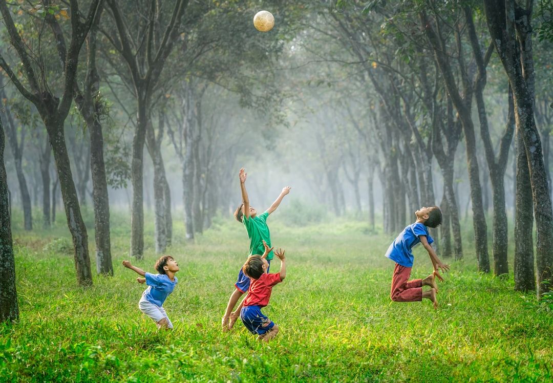 Vier Kinder spielen mit einem Ball auf einer von Bäumen gesäumten Grünfläche in Aachen. Alle vier springen aufgeregt in die Höhe, um den Ball zu erreichen. Die Szene ist üppig und grün und lässt vermuten, dass sie in einem der wunderschönen Parks der Stadt spielt.