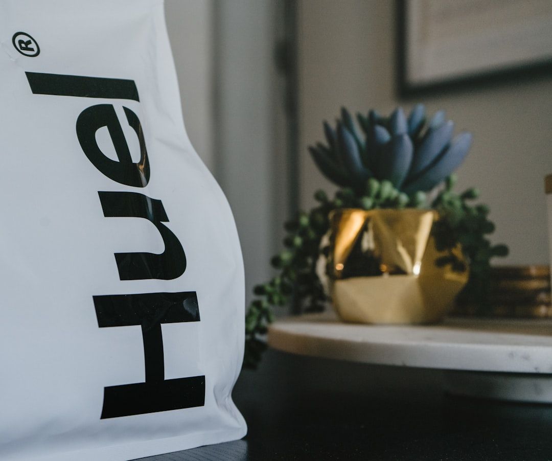 Eine weiße Huel-Lebensmittelverpackung steht auf einem Tisch neben einem goldenen Blumentopf mit einer kleinen Sukkulente und erinnert vor einem neutralen Hintergrund an die klare und minimalistische Ästhetik eines Aachener Cafés.