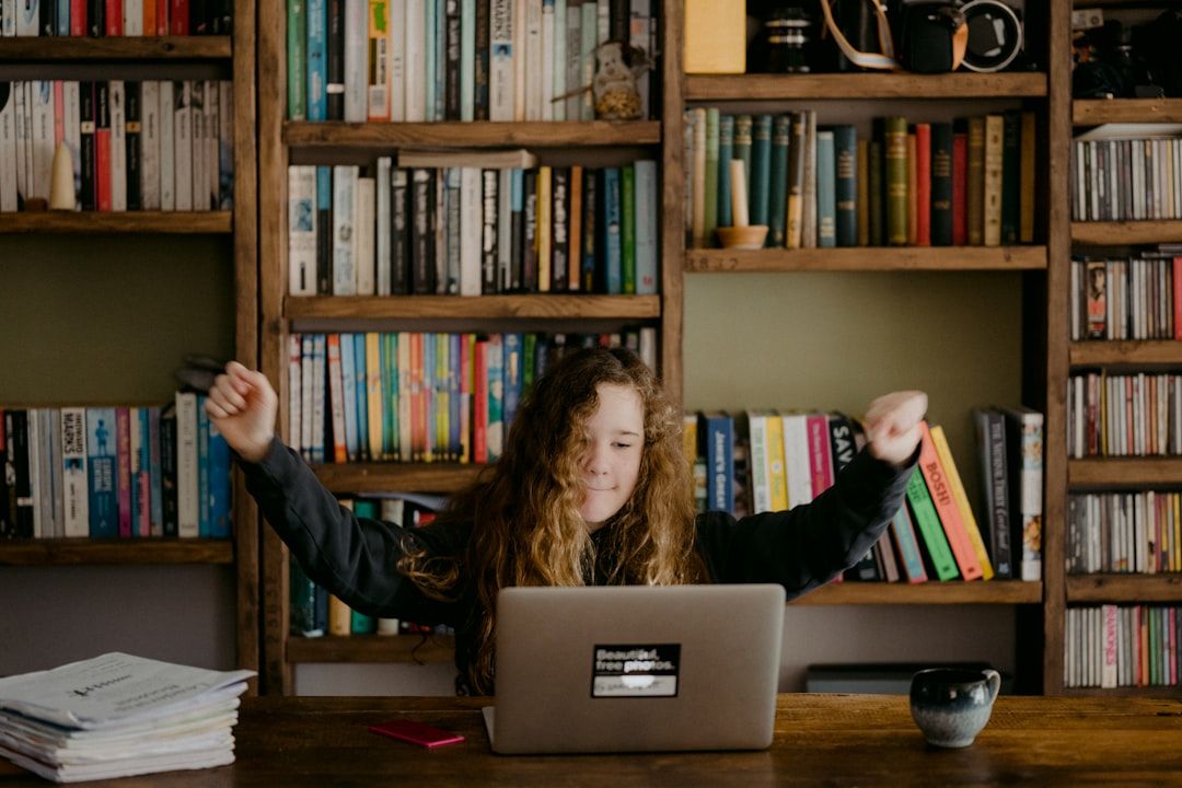 Eine Person mit langen Haaren sitzt mit einem Laptop an einem Schreibtisch und reckt aufgeregt die Arme in die Höhe. Dahinter steht ein hölzernes Bücherregal voller Bücher und verschiedener Gegenstände, das eine akademische Atmosphäre schafft, die an die angesehenen Aachener Bibliotheken erinnert. Auf dem Schreibtisch steht eine Tasse.