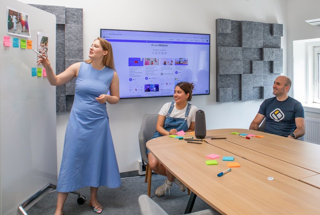 Drei Personen sitzen in einem Besprechungsraum. Eine Person steht und gestikuliert auf Haftnotizen an einem Whiteboard und bespricht Pläne für ihre bevorstehende Reise nach Aachen, während die anderen beiden an einem Tisch sitzen und vor ihnen bunte Haftnotizen und Marker liegen.