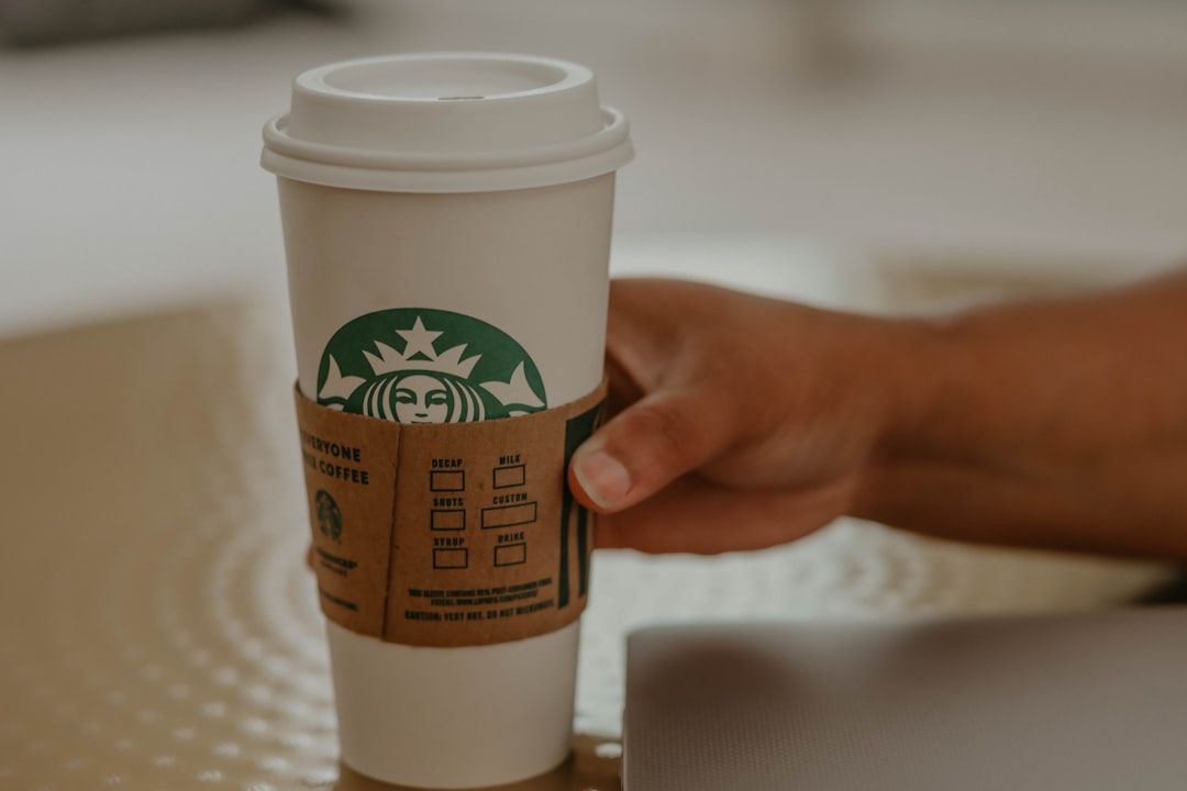 Eine Hand hält einen großen Einweg-Kaffeebecher von Starbucks mit brauner Hülle auf einem beigen Tisch und weckt Erinnerungen an gemütliche Cafés in Aachen.