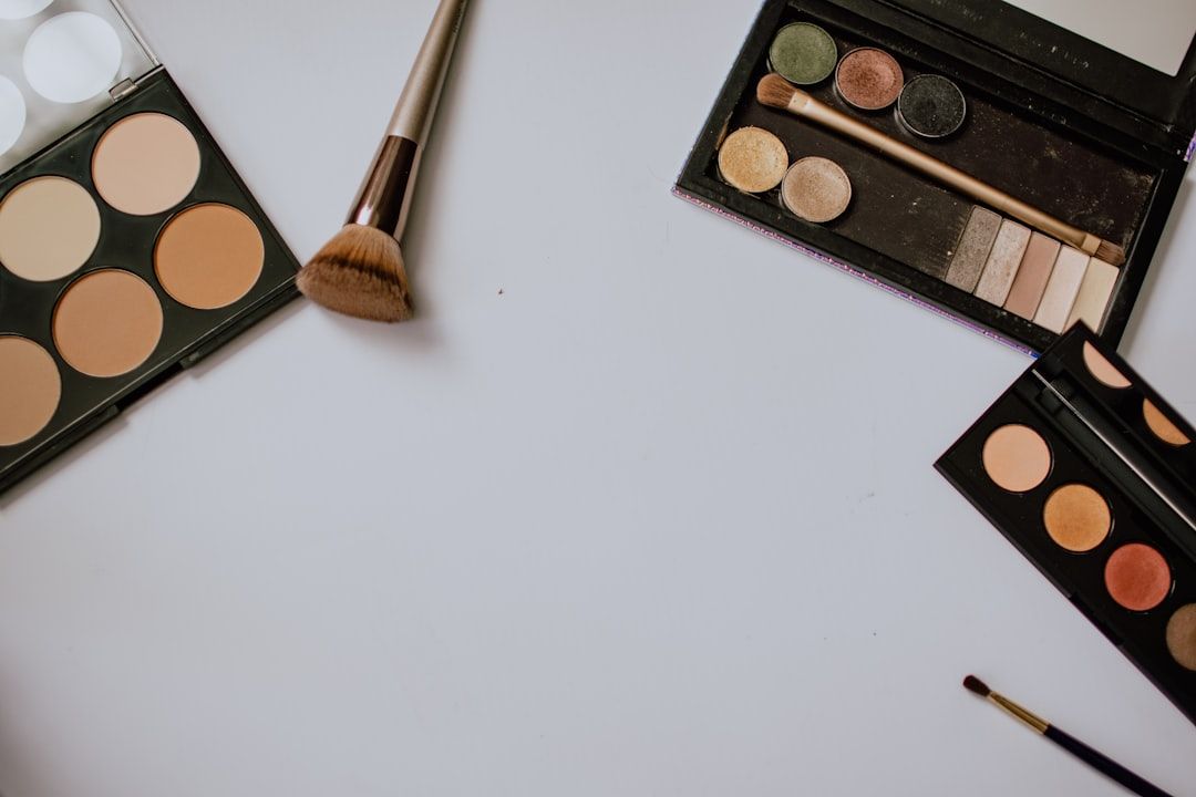 Verschiedene Make-up-Produkte, darunter Lidschattenpaletten, eine Rougepalette, ein Pinsel und ein Eyeliner-Stift, sind auf einer weißen Oberfläche angeordnet und erinnern an die elegante Schlichtheit der zeitlosen Aachener Schönheitsroutine.