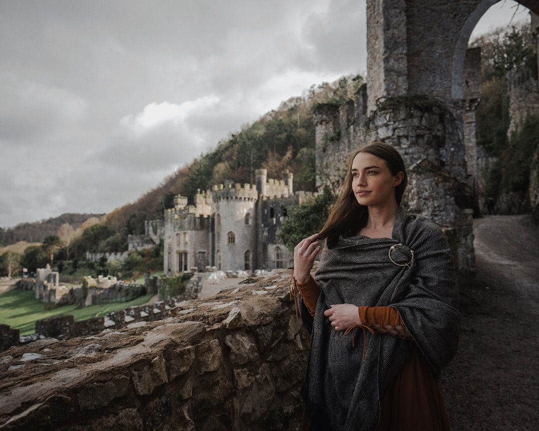 Eine Frau im mittelalterlichen Outfit steht auf einem Steinweg mit der historischen Aachener Burg und üppigen Hügeln im Hintergrund unter einem bewölkten Himmel.