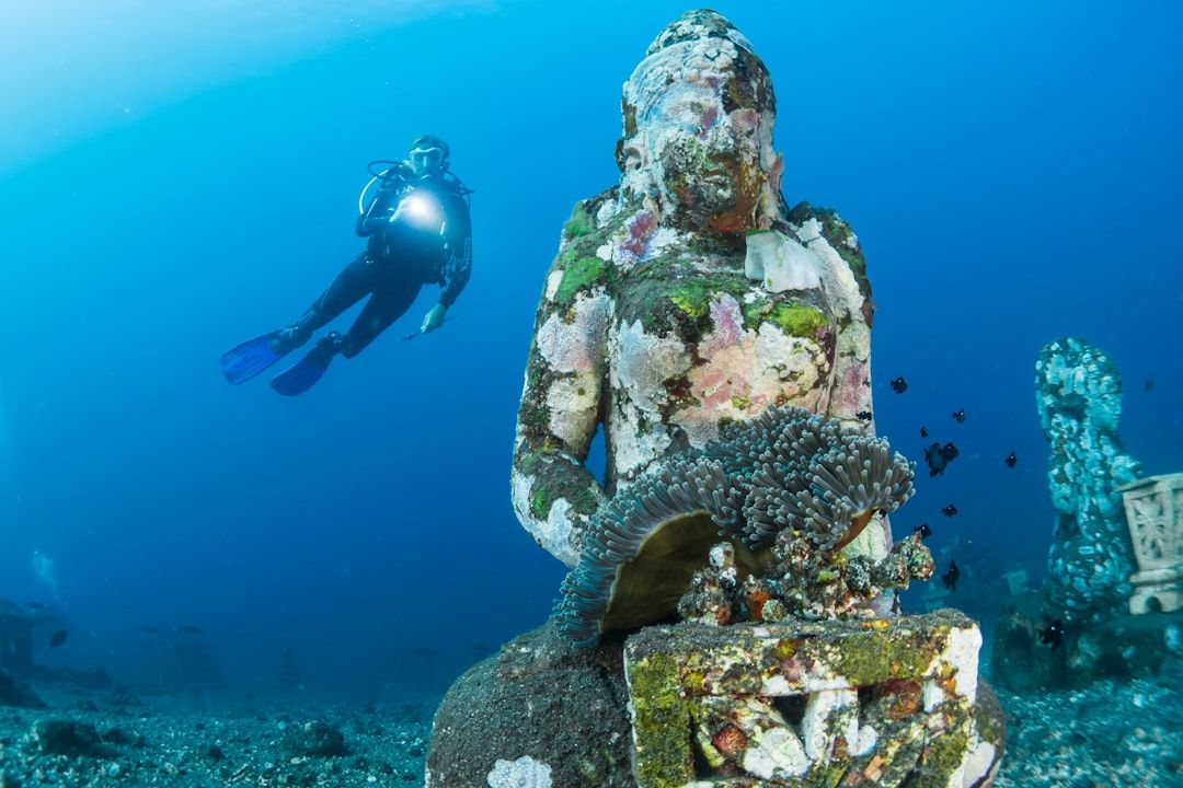 Ein Taucher schwimmt in der Nähe einer untergetauchten, algenbedeckten Statue einer sitzenden Figur, die Meereslebewesen unter Wasser hält und an die antiken Artefakte erinnert, die in Aachen gefunden wurden.