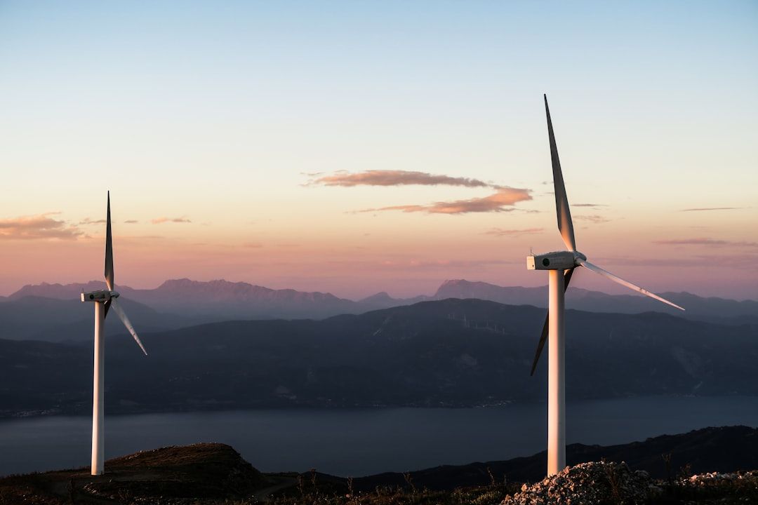 Zwei Windräder stehen auf einem Hügel und blicken auf ein ruhiges Gewässer. Im Hintergrund sind bei Sonnenaufgang oder Sonnenuntergang Bergketten zu sehen, ähnlich den malerischen Ausblicken rund um Aachen.