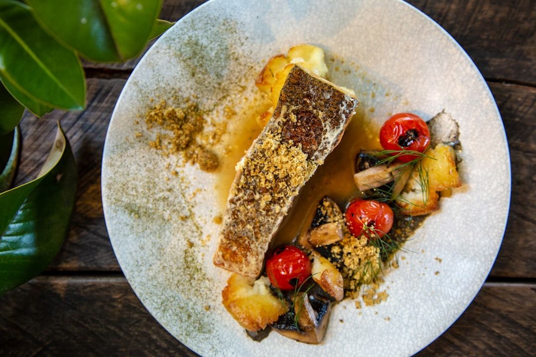 Auf einem Teller steht ein gebratenes Fischfilet mit Paniermehl, gerösteten Kirschtomaten und verschiedenen Gemüsesorten auf einem Holztisch, der an ein Aachener Café erinnert, neben einer Grünpflanze.