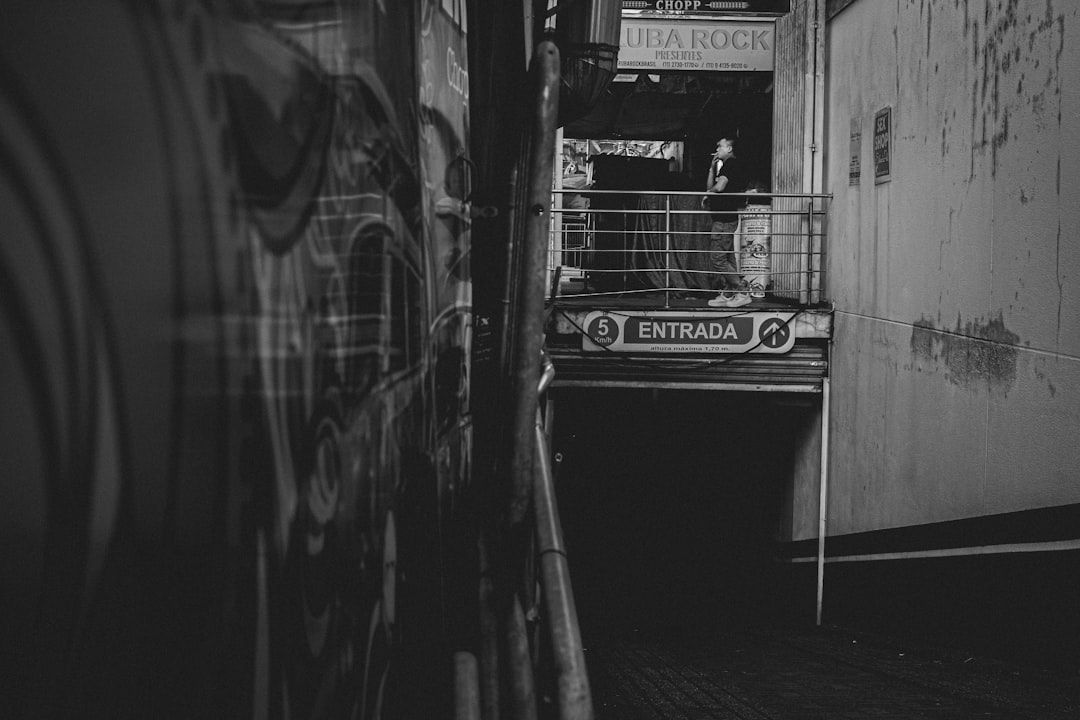 Schwarzweißfoto einer Person, die auf einem Balkon neben einem Gebäude mit der Aufschrift „Entrada“ und darüber Schildern steht. Die urbane Szene, die an die Innenstadt von Aachen erinnert, zeigt Graffiti an der Wand links.