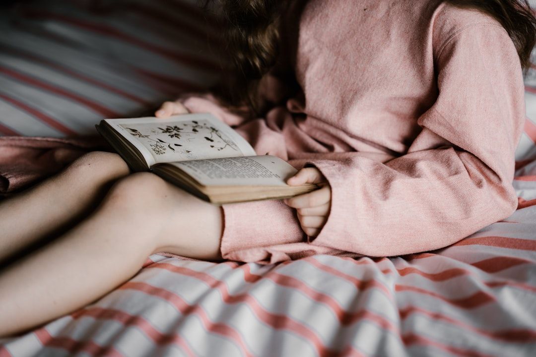 Eine Person im rosa Outfit sitzt auf einer gestreiften Bettdecke, vertieft in ein offenes Buch mit botanischen Illustrationen, und schwelgt in Erinnerungen an ihre Reise nach Aachen.
