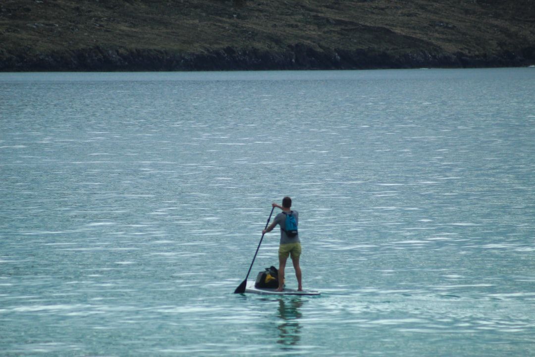 Eine Person steht und paddelt auf einem Paddleboard in ruhigen Gewässern nahe einer felsigen Küste und ruft so die friedliche Atmosphäre hervor, die an die friedliche Landschaft Aachens erinnert.