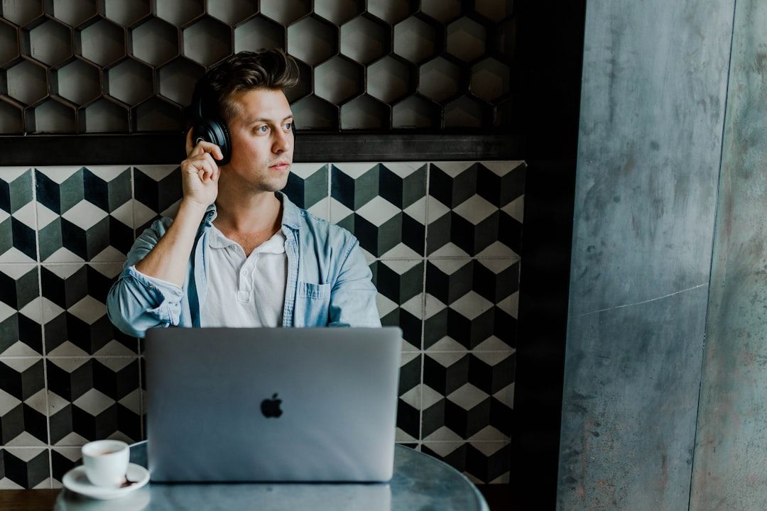 Person sitzt mit Laptop an einem Tisch, trägt Kopfhörer und blickt zur Seite. Auf dem Tisch stehen eine Tasse und eine Untertasse, im Hintergrund eine Wand mit Wabenmuster, das an die modernen Cafés Aachens erinnert.
