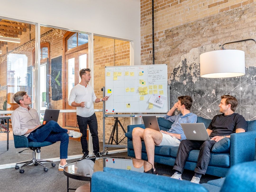 Vier Personen in einer modernen Büroumgebung; drei sitzen mit Laptops auf blauen Sofas und einer steht und präsentiert Ideen auf einem Whiteboard. Die innovative Umgebung spiegelt die Essenz von Aachens zukunftsorientiertem Ansatz zur Zusammenarbeit am Arbeitsplatz wider.