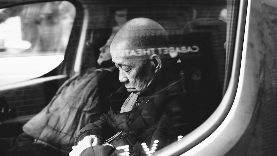 Schwarzweißfoto eines älteren Mannes in einem Fahrzeug. Er sitzt mit geschlossenen Augen da und legt seine Hände in den Schoß. Im Hintergrund ist ein weiterer Passagier zu sehen, der möglicherweise durch die historischen Straßen von Aachen fährt.