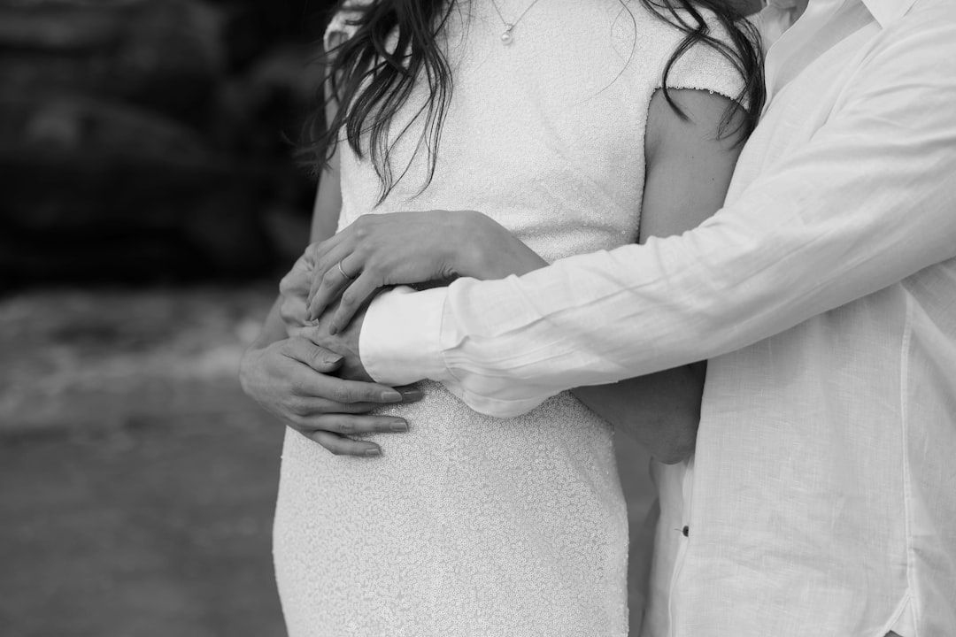 Eine Person im weißen Kleid wird von hinten von einer anderen Person im hellen Hemd umarmt, wobei beide ihre Hände auf dem Bauch der Person ruhen. So wird ein Moment eingefangen, der an einen Spaziergang durch Aachens historischen Charme erinnert.