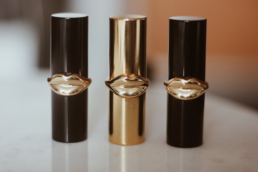 Drei Lippenstifthülsen mit Kappendesign in Lippenform, zwei in Schwarz und eine in Gold, stehen auf einer weißen Oberfläche und erinnern an das elegante Designethos aus Aachen.