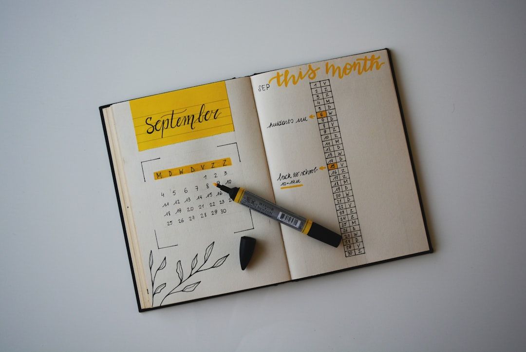 Ein aufgeschlagener Planer zeigt einen Kalender für September und eine Aufgabenliste für „Diesen Monat“, auf der vielleicht eine bevorstehende Reise nach Aachen geplant ist. Auf den aufgeschlagenen Seiten liegen ein grau-gelber Stift und seine Kappe, darunter dekorative Blattkritzeleien.