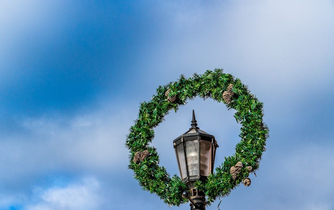 Ein grüner Kranz mit Tannenzapfen ziert einen Laternenpfahl vor einem blauen, bewölkten Himmel und erinnert an die zauberhaften Straßen von Aachen.