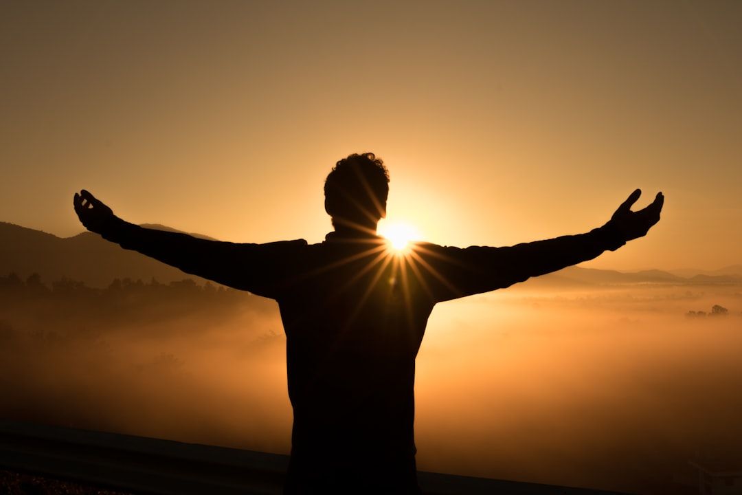 Eine Person steht mit ausgestreckten Armen dem Sonnenaufgang entgegen, mit Bergen und Nebel im Hintergrund, was an die heitere Schönheit Aachens erinnert.