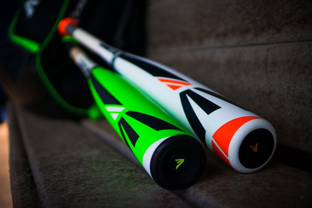 Zwei Baseballschläger, einer grün-schwarz und einer orange-schwarz, liegen auf einer Holzbank in Aachen, im Hintergrund ist eine Sporttasche zu sehen.