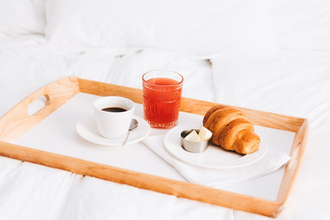 Auf einem Bett steht ein Holztablett mit einer Tasse Kaffee, einem Glas Saft, einem Croissant und einem Schälchen mit Butterstückchen, das an ein elegantes Aachener Frühstück erinnert.