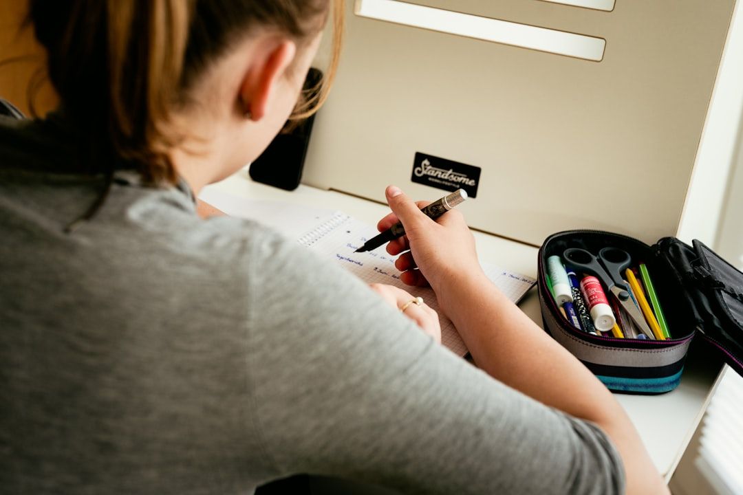 Person sitzt an einem Schreibtisch in Aachen und schreibt mit einem Stift in ein Notizbuch. Auf dem Schreibtisch ist ein Federmäppchen mit verschiedenen Schreibwaren zu sehen.