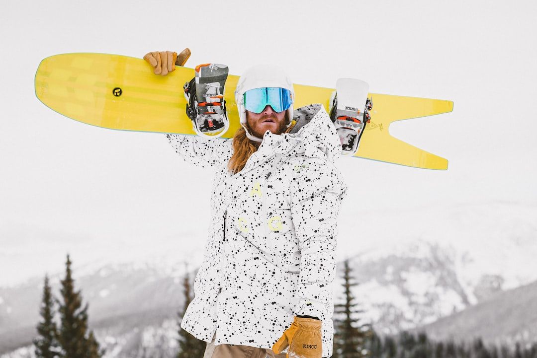 Eine Person mit gemusterter weißer Jacke, Skibrille und Helm hält ein gelbes Snowboard über der Schulter. Im Hintergrund sind schneebedeckte Berge und Kiefern zu sehen, die an die malerische Landschaft bei Aachen erinnern.