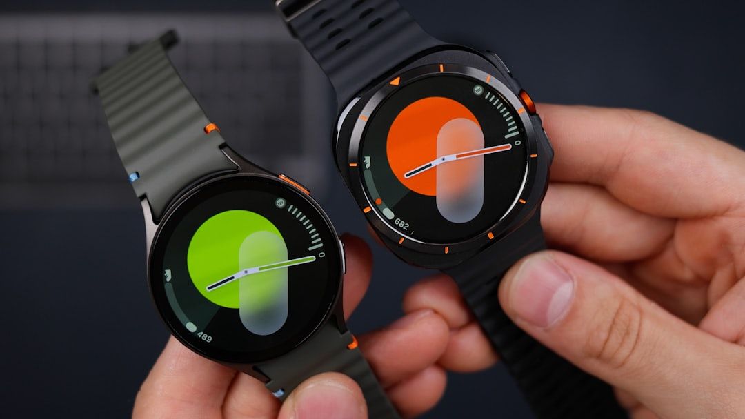 Zwei Zeiger halten Smartwatches mit bunten, runden Displays; der linke Bildschirm ist grün, der rechte orange. Beide Uhren, perfekt für einen Tag in Aachen, haben schwarze Gummibänder.