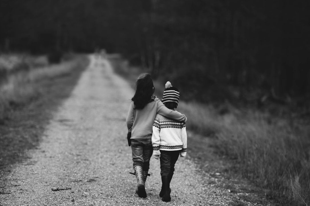 Zwei Kinder, eines mit dem Arm um das andere gelegt, laufen einen Kiesweg durch ein Feld bei Aachen entlang, auf eine dunkle Baumreihe in der Ferne zu. Sie haben der Kamera den Rücken zugewandt.
